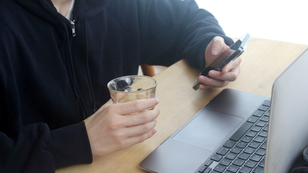 PCの前で飲み物を片手にスマホを操作する手元