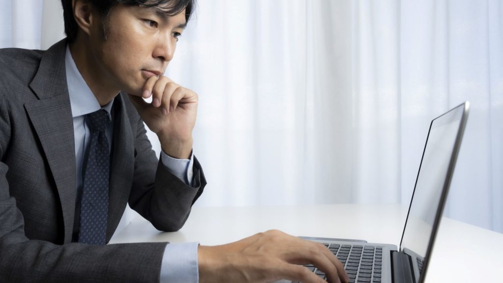 PC画面を見つつ顎に手を当てて悩む男性