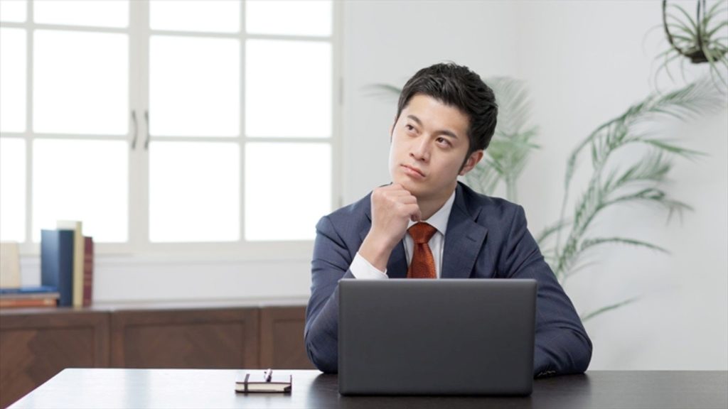 PCの前で肘をついて斜め上を見つつ悩む男性